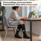 Kobieta w profilu przy biurku z laptopem w zimowym świetle z okna, na nogach podgrzewane podkolanówki, stopy na drewnianej podłodze